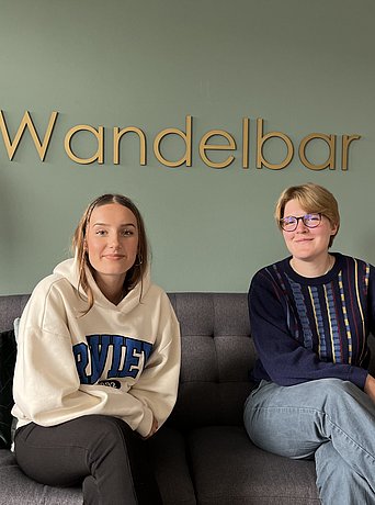 Die Auszubildenden Leni Röver und Tarja Hesse sitzen auf einer Couch vor dem Hintergrund der Wandelbar.