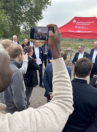 Ein Gruppe steht auf einer Freifläche, im Hintergrund ist ein rotes Pavillondach zu sehen. Im Vordergrund macht ein Mann ein Foto mit seinem Smartphone.