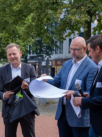 Eine Gruppe im Gespräch, ein Mann in der rechten Bildmitte hat Baupläne in der Hand.