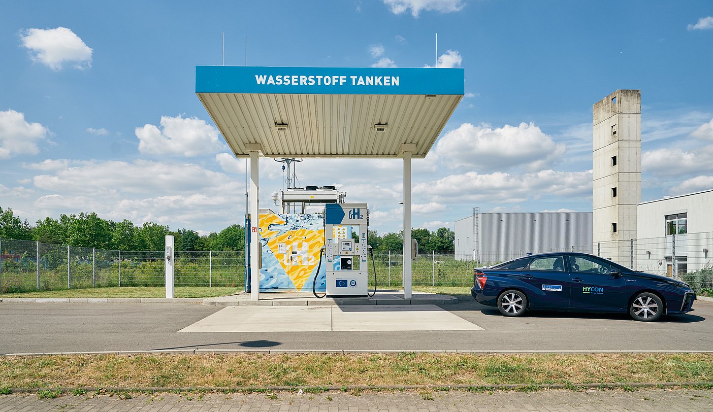 A hydrogen filling station is depicted with the lettering Refuel hydrogen. The filling station is left by a refueled hydrogen car.