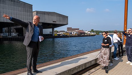 Ein Mann steht an einem Kanal und zeigt auf etwas, eine Gruppe hört im zu.