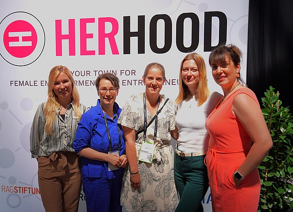 Fünf Frauen die vor dem HerHood Logo in die Kamera lächeln.